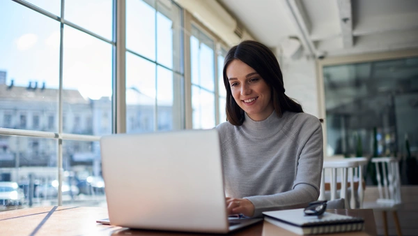 Eine junge Frau sitzt am Fenster und blickt in ihren Laptop